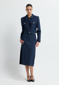 Tmavě modrý dvoudílný outfit s krátkým sakem s velkými knoflíky, dvěma předními kapsami a kolenní sukní s bočním rozparkem.