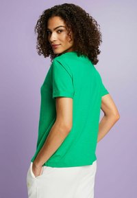 Camiseta de manga corta verde hecha de tejido ligero, con un corte clásico, cuello redondo y mangas enrolladas, exhibida sobre un fondo púrpura.