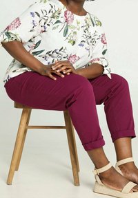 Persoon die een witte bloes met bloemenprint, geborduurde bordeauxrode opgerolde broek en beige geweven sandalen draagt, zittend op een houten kruk tegen een effen achtergrond.