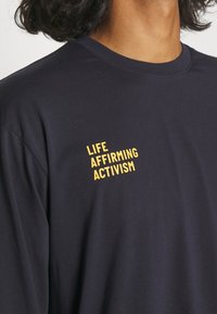 En person som bär en svart tröja med gul text på bröstet som säger "LIVSBEKRÄFTANDE AKTIVISM", som visar överkroppen och nacken.