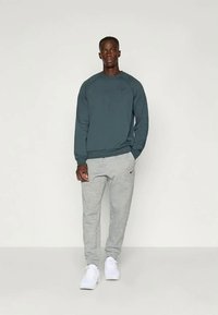 Felpa a maniche lunghe di colore verde scuro con maniche raglan e pantaloni della tuta grigi in tessuto testurizzato. L'outfit è completato da sneakers bianche.