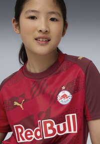 Camiseta de fútbol roja con tonos rojos y burdeos, patrón texturizado, logo de Puma, escudo del FC Salzburg y "Red Bull" en texto blanco destacado.