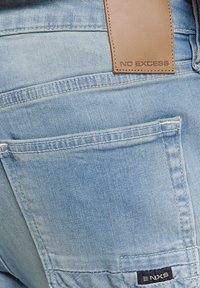 Lichtblauwe spijkerbroek van denim met een leren label met de tekst "NO EXCESS" op de achterkant, een enkele achterzak en zichtbare stikseldetails.