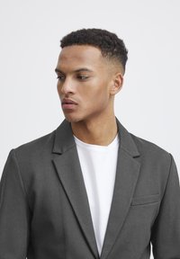 Dunkelgrauer Blazer mit einer einzelnen Brusttasche, Reverskragen und glatter Textur, kombiniert mit einem weißen Rundhals-T-Shirt darunter.