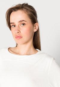 Top leve e branco com decote arredondado e textura suave, usado por uma pessoa com cabelo comprido, destacando as feições do rosto.