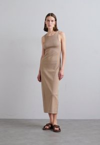 Abito maxi beige senza maniche realizzato in tessuto a costine con una silhouette aderente. Abbinato a sandali marroni annodati. Sfondo neutro.