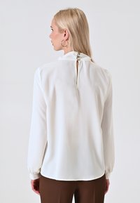Weiße Bluse mit langen Ärmeln, hohem Kragen, Schlüsselbereichsverschluss hinten und Knopfdetails. Glattes Material mit sanftem Glanz, leicht abgerundeter Saum.