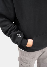 Felpa nera con finitura testurizzata, vestibilità oversized e polsini a coste. Presenta un piccolo logo geometrico bianco sulla manica sinistra.
