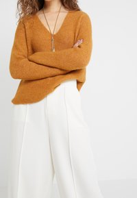 Musztardowy sweter dzianinowy z głębokim dekoltem w serek, prążkowaną fakturą i długimi rękawami, w połączeniu z białymi spodniami o szerokich nogawkach. Długi naszyjnik z zawieszką.