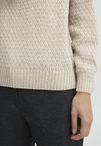 Maglione lavorato a maglia color crema con un motivo testurizzato, orlo e polsini a costine. Abbinato a pantaloni leggeri grigio scuro.