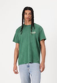Groene katoenen t-shirt met een cirkelvormig logo op de borst en versleten details. Losse fit en korte mouwen. Gecombineerd met lichtblauwe jeans.