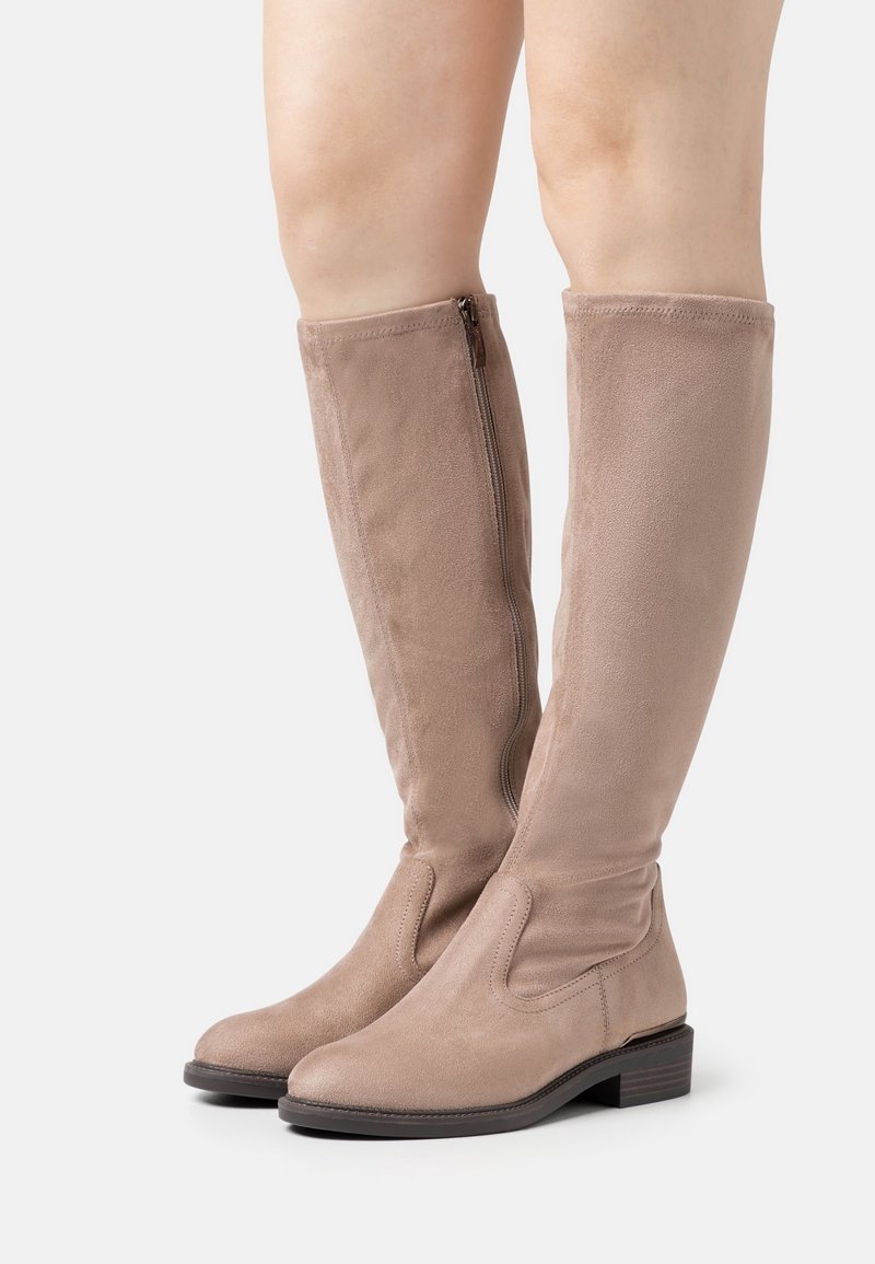 Bottes en daim beige montant jusqu'au genou avec fermeture éclair latérale, talon bas empilé, portées sur des jambes nues sur fond blanc.