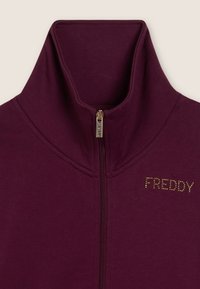 Garment con collo alto color bordeaux con zip e tirazip dorato, con logo "FREDDY" in piccoli chiodini dorati sul petto.