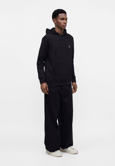 Homme portant un sweat à capuche noir, un pantalon large noir et des baskets blanches, debout devant un fond blanc uni.