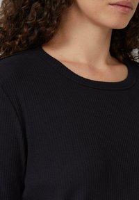 Zwart geribbelde top met een ronde halslijn. Zachte textuur, aansluitend ontwerp dat de nek en schouderpartij accentueert. Verstelbaar stiksel detail.