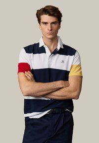 Jeune homme les bras croisés portant un polo rayé bleu marine, blanc, rouge et jaune, ainsi qu'un pantalon foncé, sur un fond uni.