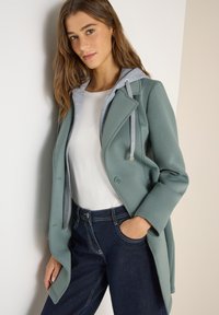Jeune femme aux longs cheveux portant un manteau vert superposé à un sweat à capuche gris et un t-shirt blanc, associée à un jean bleu foncé, les mains dans les poches.