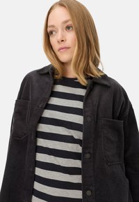 Schwarze gerippte Cordjacke mit Kragen und Brusttasche, über einem navy- und grau gestreiften Hemd getragen. Weicher Stoff, lässige Passform.