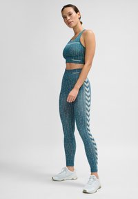 En petrolfärgad sport-bh och högmidjade leggings har vertikala ränder och chevrondetaljer. Tyget verkar elastiskt och mjukt. Vita träningsskor fullända looken.