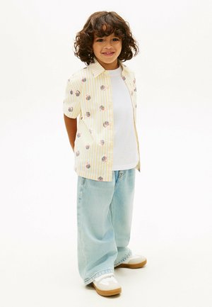 Smilende lille barn med krøllet hår iført en gul stribet skjorte med mønstre, hvid t-shirt, lyseblå jeans og hvide sneakers, stående.