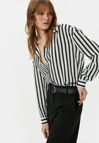 Blusa a rayas en blanco y negro con un profundo escote en V, botones al frente y mangas largas, combinada con pantalones negros de tiro alto y un cinturón.