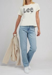 Camiseta de manga corta crema con logo negro "Lee", combinada con jeans rectos de mezclilla azul claro y zapatillas blancas, sosteniendo una chaqueta de rayas beige.