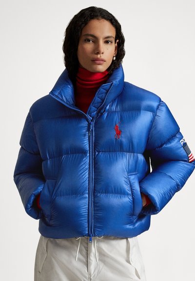 Blue puffer jacket with a glossy finish, featuring a high collar, quilted design, and red embroidered logo. Includes zip closure and flag accents.