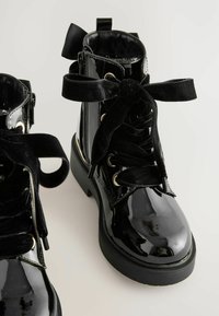 Botas de tobillo de charol negro con cordones de terciopelo, ojales dorados y un tacón grueso. Presentan un forro textil y una lengüeta de agarre.