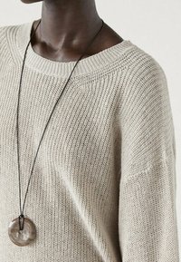 Pull en maille beige avec un décolleté large, présentant un motif texturé. Un collier avec un pendentif circulaire suspendu à un cordon noir.