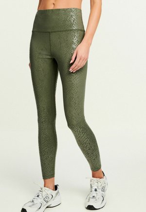Donna che indossa leggings high-waisted verde oliva con un sottile motivo a serpente e scarpe sportive bianche, in piedi con una mano rilassata.