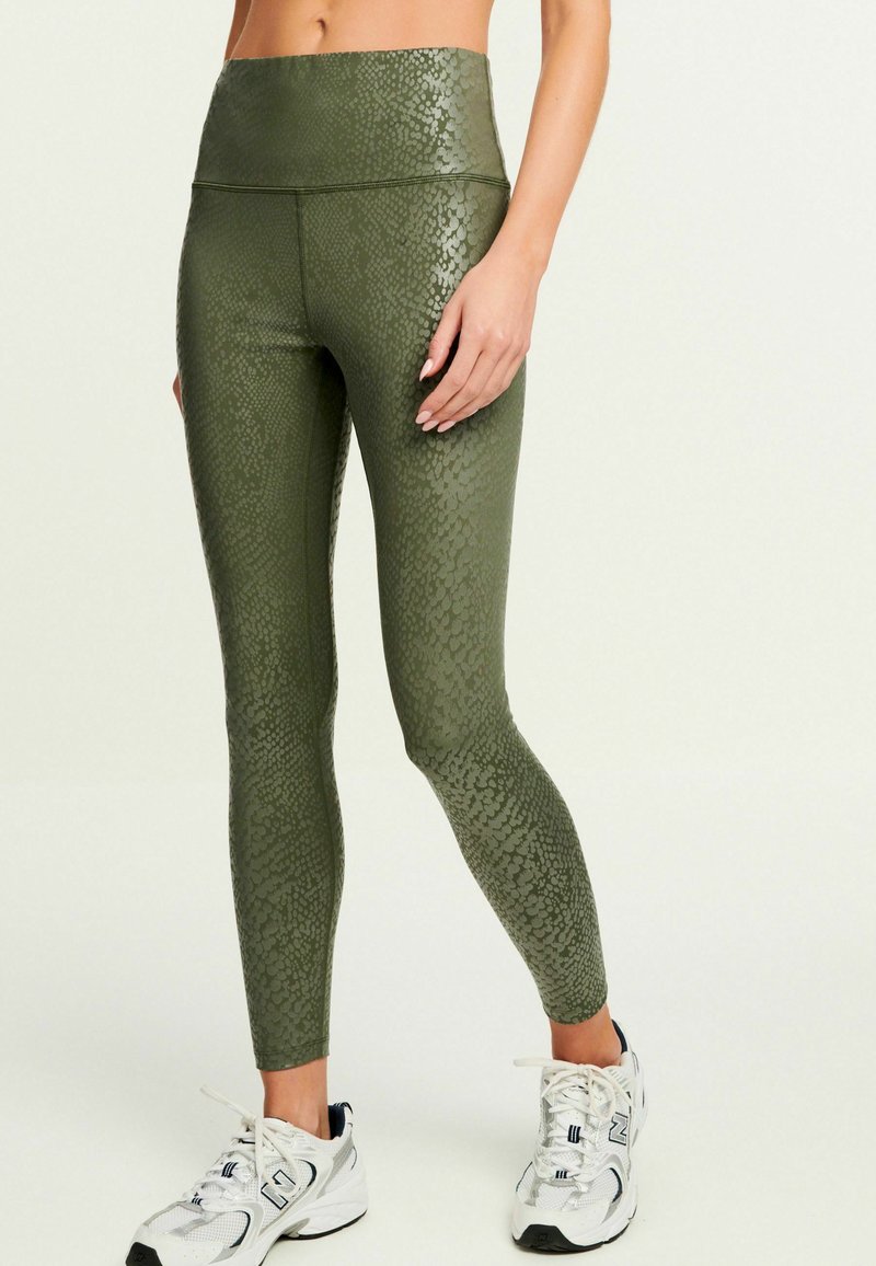 Kvinna iförd högmidjade olivgröna leggings med subtilt ormskinnsmönster och vita träningsskor, står med en hand avslappnad.