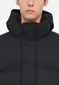 Sort pufferjakke med høj krave og snor, med en glat, mat finish og horisontalt kviltet design til isolering.