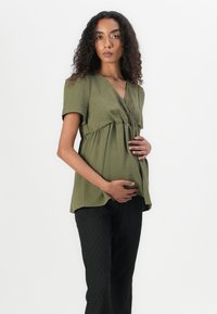 Haut de maternité vert olive à manches courtes avec un décolleté en V, taille froncée et coupe ample, associé à un pantalon large noir texturé.