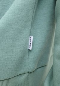 Primer plano de tela verde suave que muestra una etiqueta blanca de la marca Jack & Jones cosida en la costura de una prenda de punto.