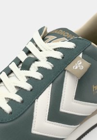Sneaker verde e bianco con tessuto testurizzato, design a zigzag a contrasto e lacci bianchi prominenti. Presenta un'etichetta con logo beige. Suola nera.
