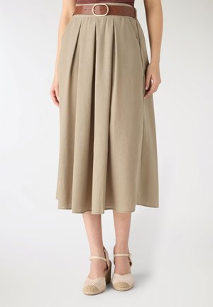 Donna che indossa una gonna midi plissettata beige con tasche laterali, una cintura marrone e scarpe con zeppa beige con cinturino alla caviglia, in piedi davanti a uno sfondo semplice.