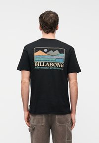 Άνδρας που φοράει μαύρο t-shirt με πολύχρωμο γραφικό βουνό και σελήνη και το κείμενο "BILLABONG Adventure division" στην πλάτη, συνδυασμένο με καφέ παντελόνι cargo.