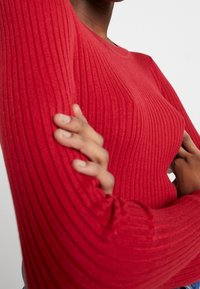 Pull rouge à manches longues côtelé avec un col rond, un tissu texturé et une coupe ajustée. Gros plan sur le bras et la position de la main.