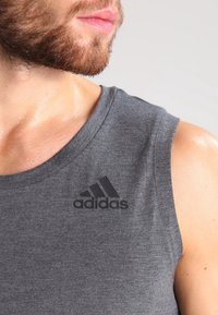 Vousatý muž v šedém tričku bez rukávů značky Adidas, ukazující svůj krk, rty a pravé rameno na jednobarevném pozadí.