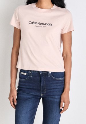Frau trägt ein hellrosa Calvin Klein Jeans T-Shirt und dunkelblaue Jeans, steht vor einem einfarbigen Hintergrund.