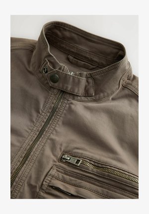Giacca in tessuto leggero marrone con colletto alto, diverse tasche con zip e finitura testurizzata. La zip è in metallo argentato.