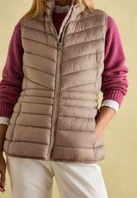 Beige gepolsterte Weste mit vertikaler Naht, Reißverschluss und Seitentaschen, über einem burdeauxfarbenen Pullover mit gerippten Bündchen und Spitzendetails getragen.