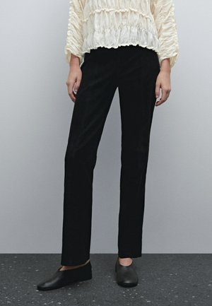 Vrouw die zwarte broek met rechte pijpen, zwarte instapschoenen en een witte blouse met lange mouwen en textuur draagt, staand op een donkergevlekt vloer.