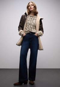 Brun fårskinnsrock med krämfärgad foder, mönstrad beige tröja och mörkblå vida jeans. Bruna ankelboots fullbordar outfiten.