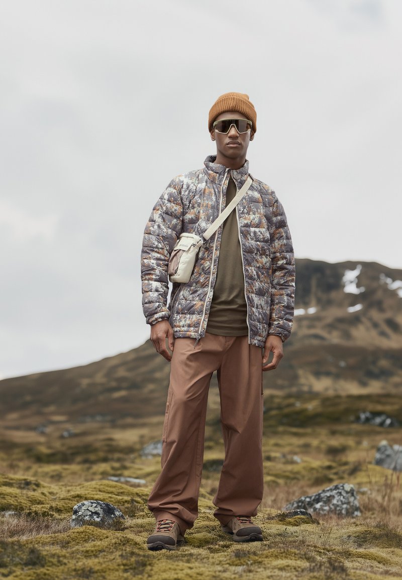 Puchowa kurtka z wielokolorowym wzorem, oliwkowa koszula, brązowe spodnie, beżowa czapka i szare buty; mała torebka noszona na ciele.