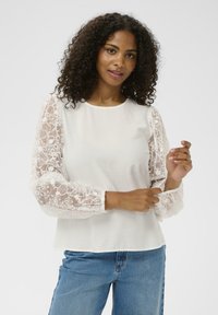 Haut blanc avec manches bouffantes en dentelle transparente, ornées de broderies florales. Texture lisse, encolure ronde et coupe décontractée. Associé à un jean bleu.