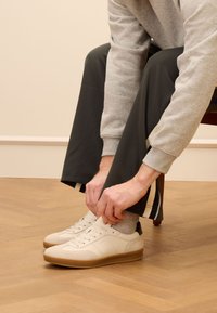 Sneaker bianche con una finitura liscia, suola in gomma e tallone nero. Maglione grigio e pantaloni scuri, seduto con attenzione alla calzatura.