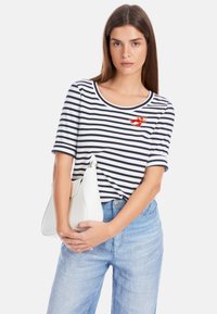 Striped t-shirt in navy and white with short sleeves, featuring a red lobster design. Paired with light blue denim and a white bag.