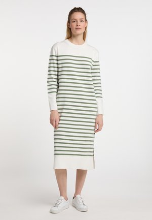 Jonge vrouw die een witte midi-jurk met groene horizontale strepen en witte sneakers draagt, staand tegen een effen achtergrond.