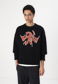 Sort strikket sweater med et iøjnefaldende abstrakt design i rød og hvid. Har en rund halsudskæring og ribbede manchetter for en afslappet pasform.